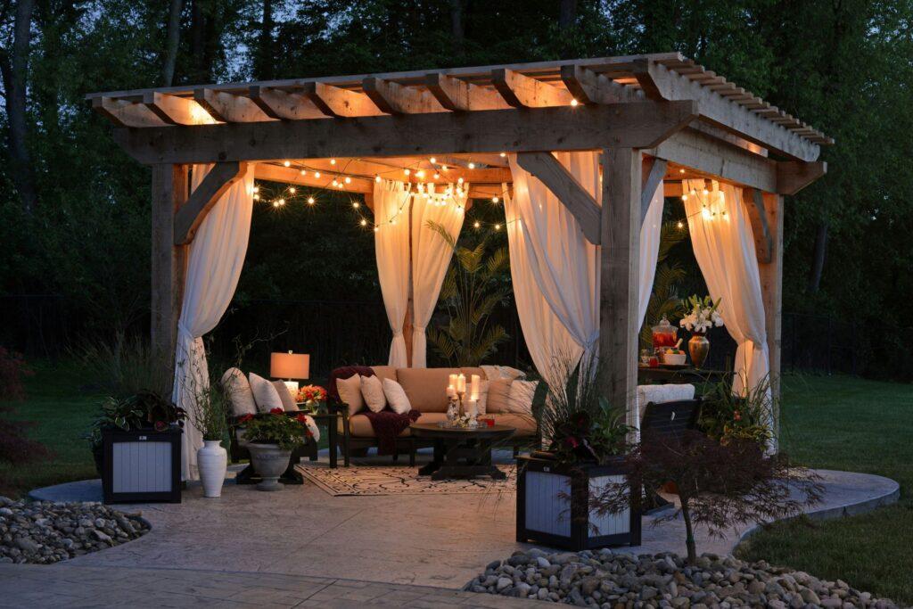 Eine gemütliche Lounge unter einer Pergola, dekoriert mit Vorhängen, Lichterketten und Pflanzen, ideal für ein Abend-Gartenfest.